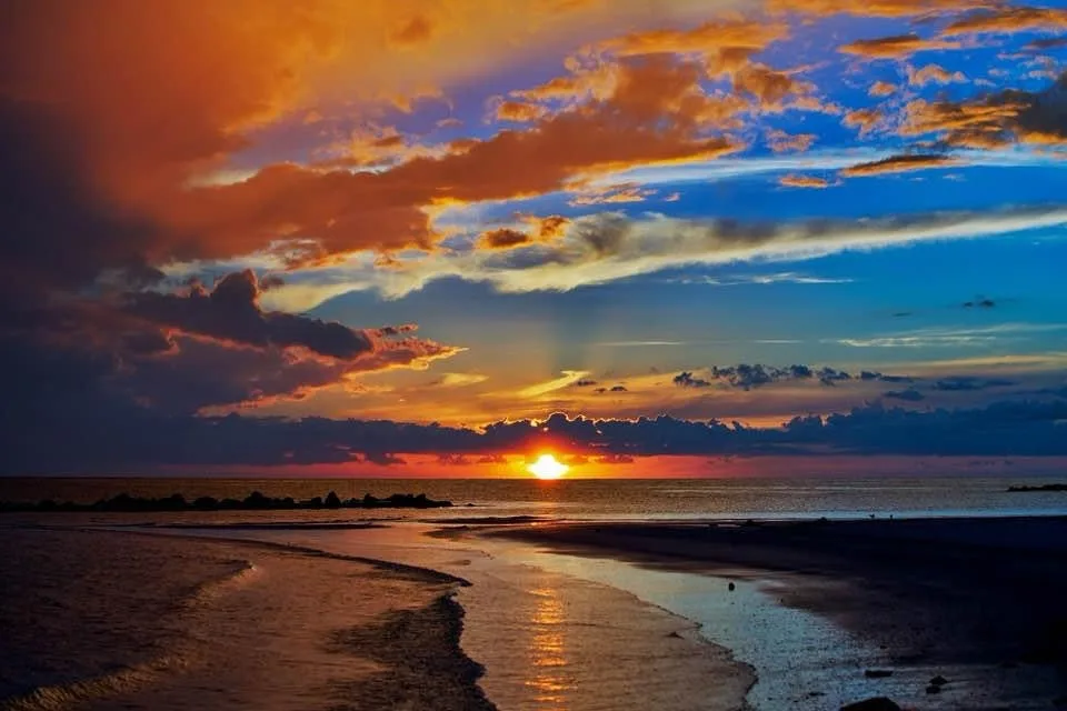 A vibrant sunset over a beach with orange, red, and blue clouds, the sun low on the horizon reflecting on wet sand. Overlaid…