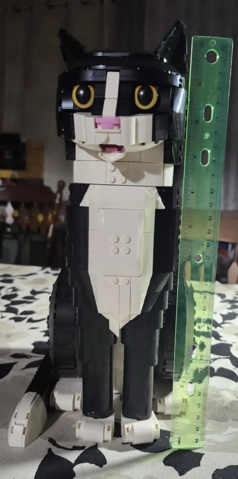 a large, black-and-white lego cat model sits upright next to a green ruler, which shows its height as 26 cm. The cat has…