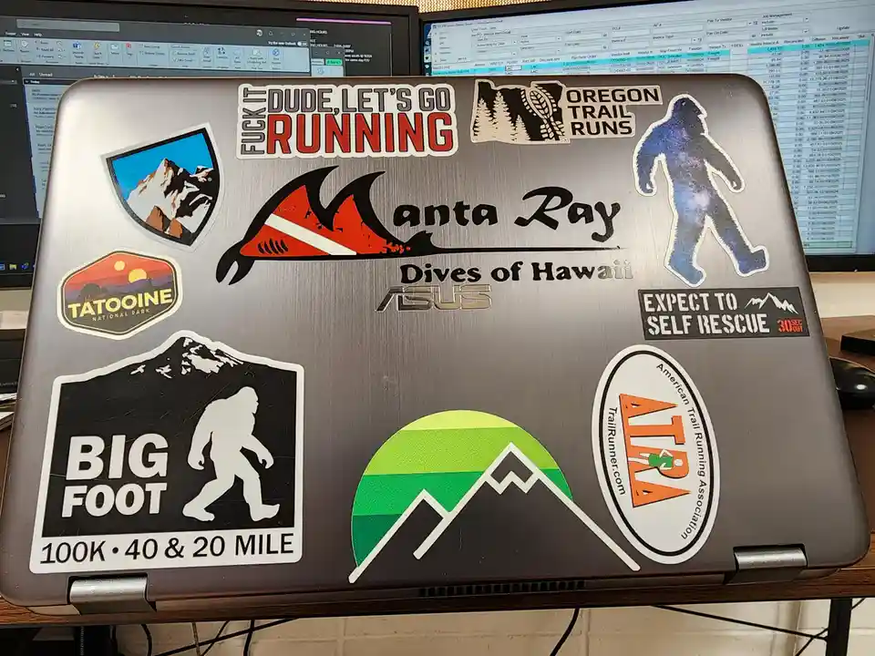 The image shows the lid of a silver ASUS laptop covered in various stickers, including 'FUCK IT DUDE, LET'S GO RUNNING'…