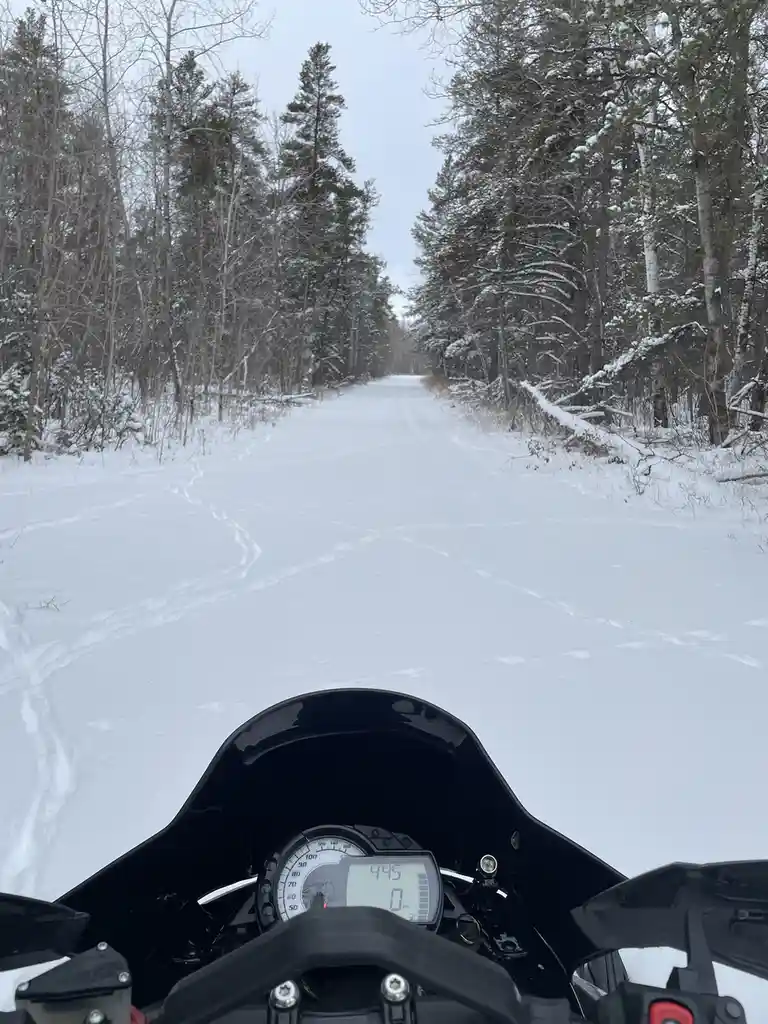 A first-person view from a motorcycle on a snow-covered road flanked by snow-laden trees, with the bike's speedometer…