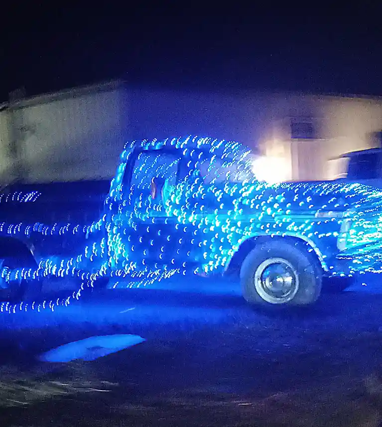 A vintage pickup truck is covered in bright blue string lights, captured at night with motion blur, making the lights appear…