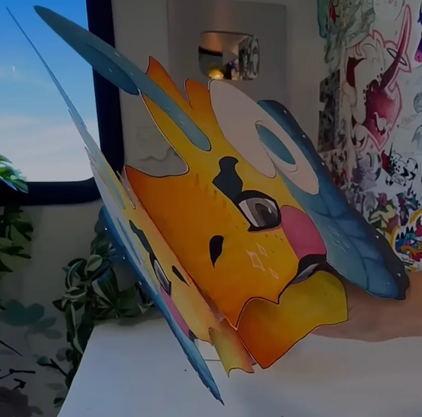 A hand holds a large, colorful, 3D paper cutout of a stylized cartoon dragon with orange, yellow, and blue wings and horns…