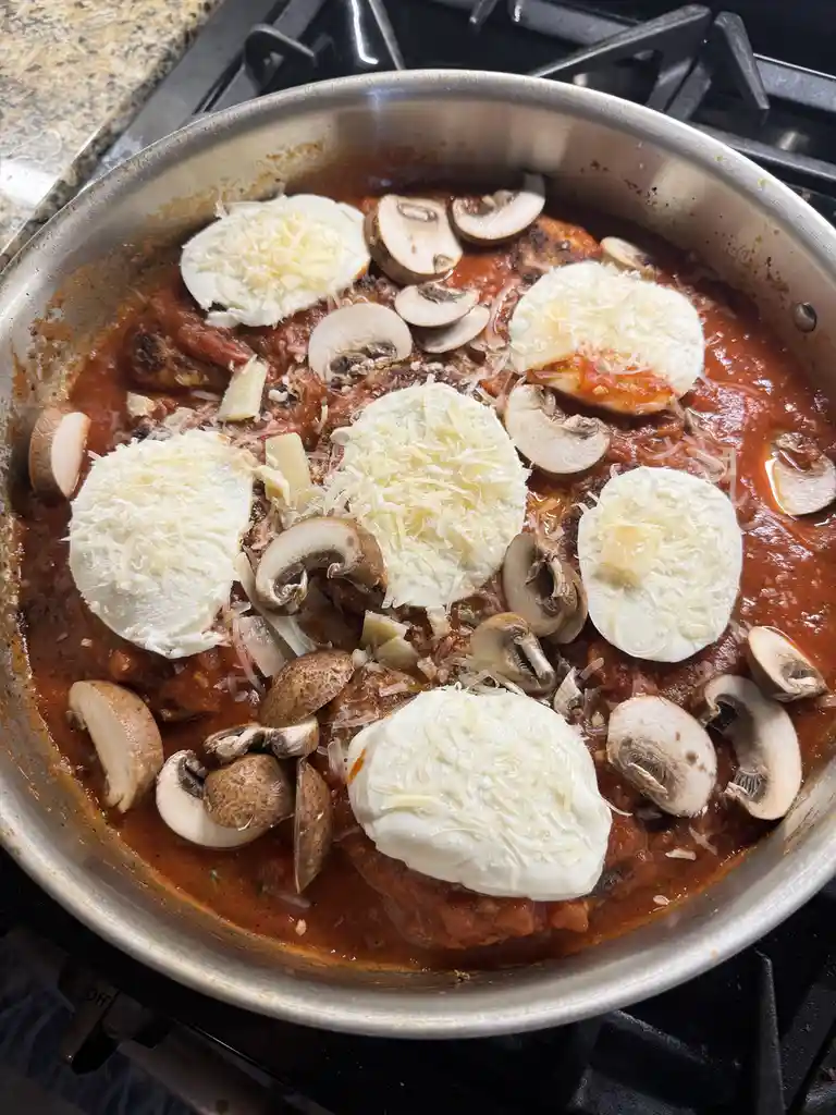 A stainless steel skillet on a stovetop contains chicken pieces in tomato sauce, topped with sliced mushrooms and multiple…