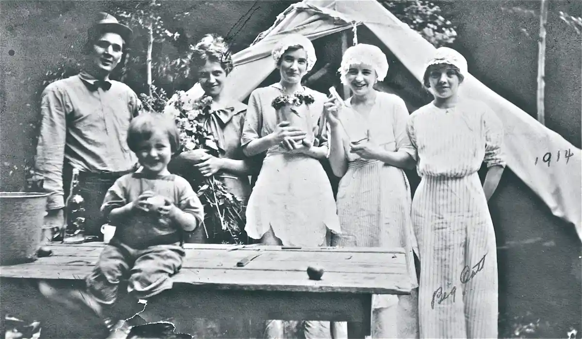 A vintage 1914 black-and-white photo shows a group of six people — a man, a young child, and four women — posing outdoors in…