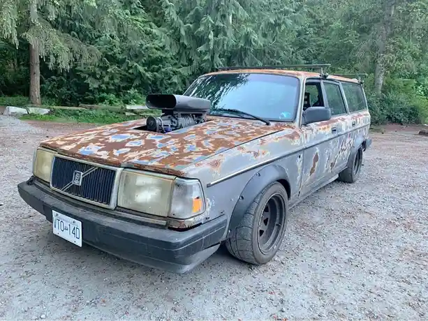 A heavily rusted, modified Volvo station wagon with a supercharger mounted on its hood, parked on gravel with trees in the…