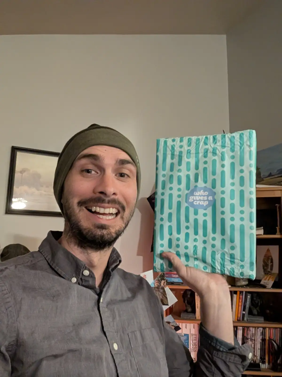 A man in a grey button-up shirt and olive green beanie smiles broadly at the camera while holding up a light blue bag with…