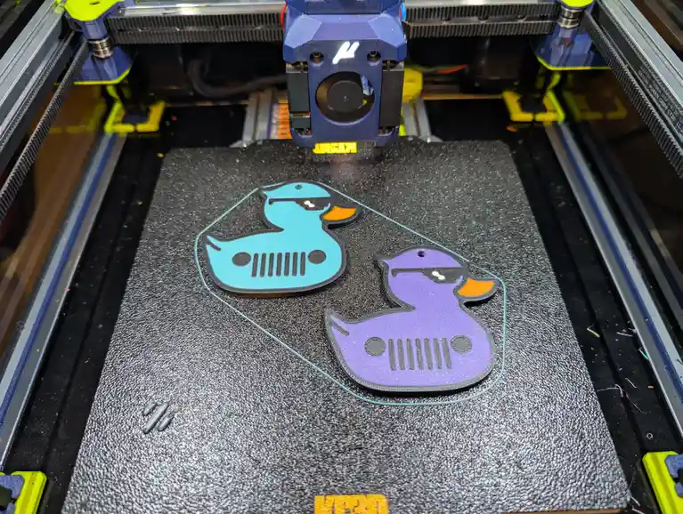 A 3d printer is actively printing two duck-shaped objects on its build plate, one light blue and one purple, both designed…