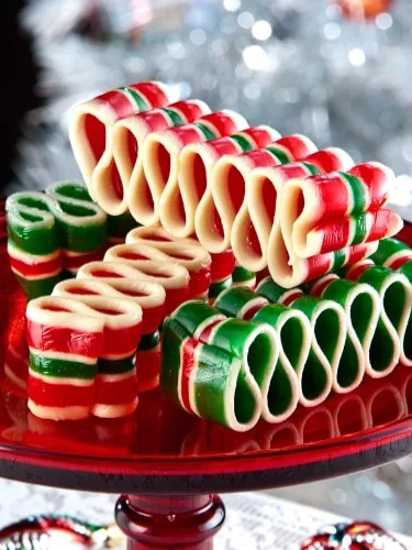 A festive display of colorful, striped, candy cane-shaped gummy candies arranged in a zigzag pattern on a red glass cake…