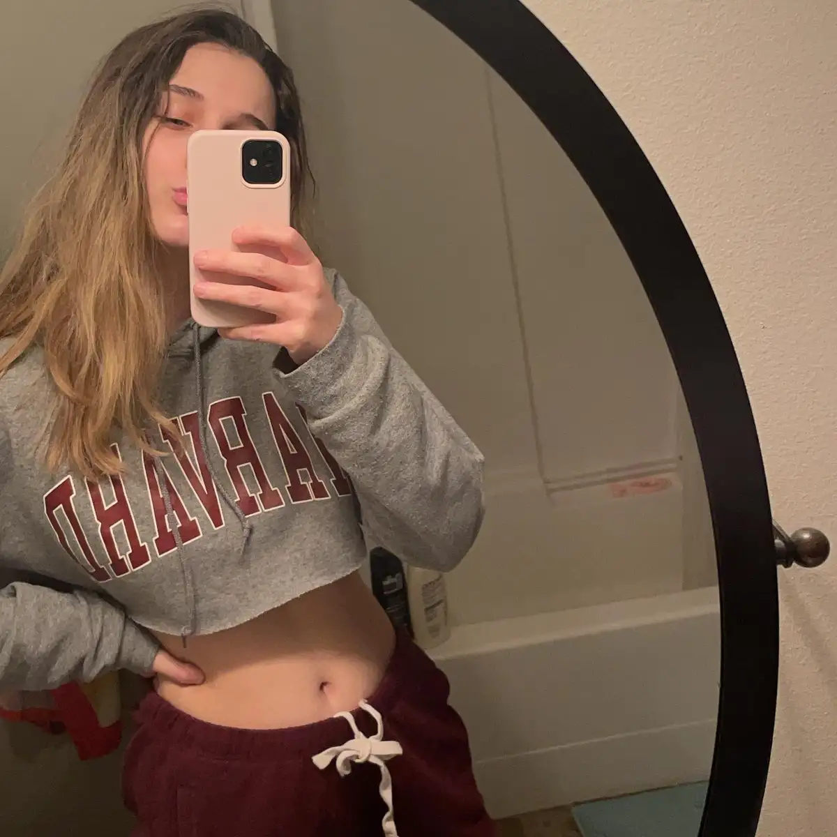 A young woman with long brown hair takes a mirror selfie in a bathroom, wearing a cropped grey hoodie with 'HARVARD' in red…