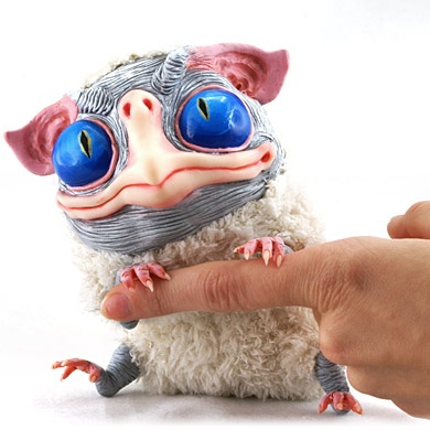 A small, fluffy toy resembling a Gremlin with large blue eyes and pink ears is being held by a human finger pointing at its…