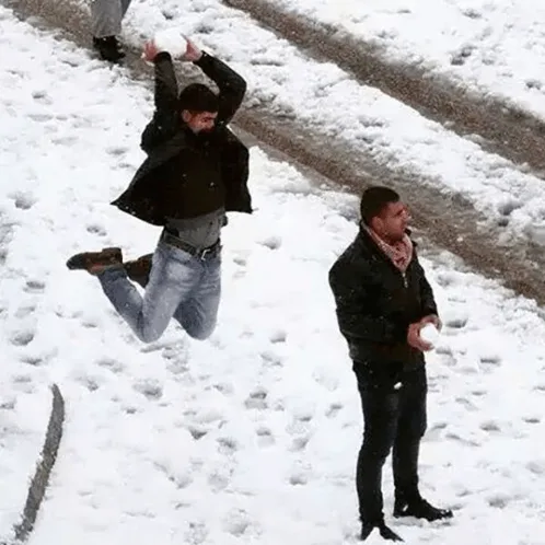 A man in a black jacket and jeans leaps mid-air in a snowy area, arms raised as if throwing a snowball upward, while another…