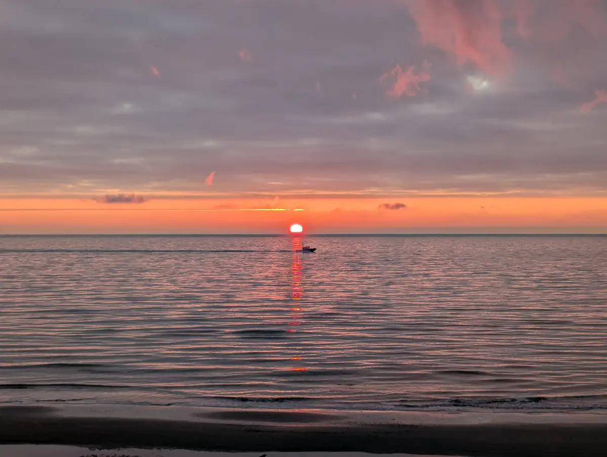 A serene sunset over the ocean with the sun low on the horizon, casting a long, shimmering reflection across the water. A…