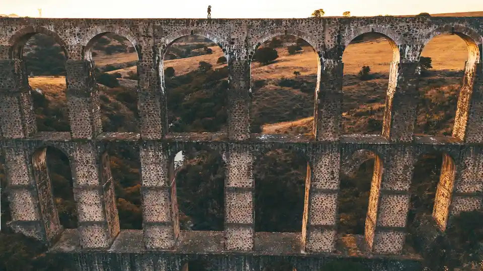 Ancient arches stretch across the landscape, a rust-hued silhouette against a blaze of golden light. Verdant foliage, a…