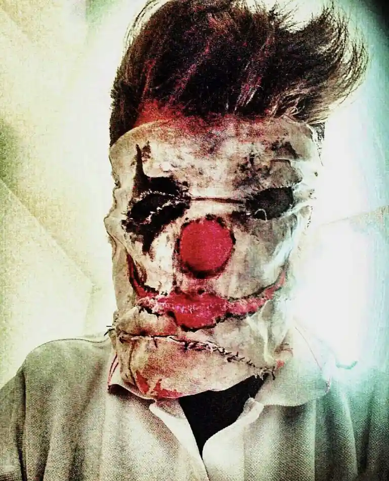 A person with dark hair wears a tattered, stitched clown mask featuring a red nose, black eye patches, and red lips, over a…