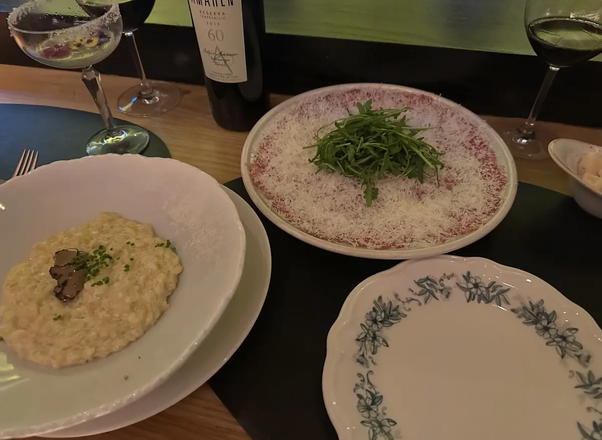 A dining table displays a bowl of truffle risotto, a plate of beef carpaccio topped with arugula and parmesan, and an empty…