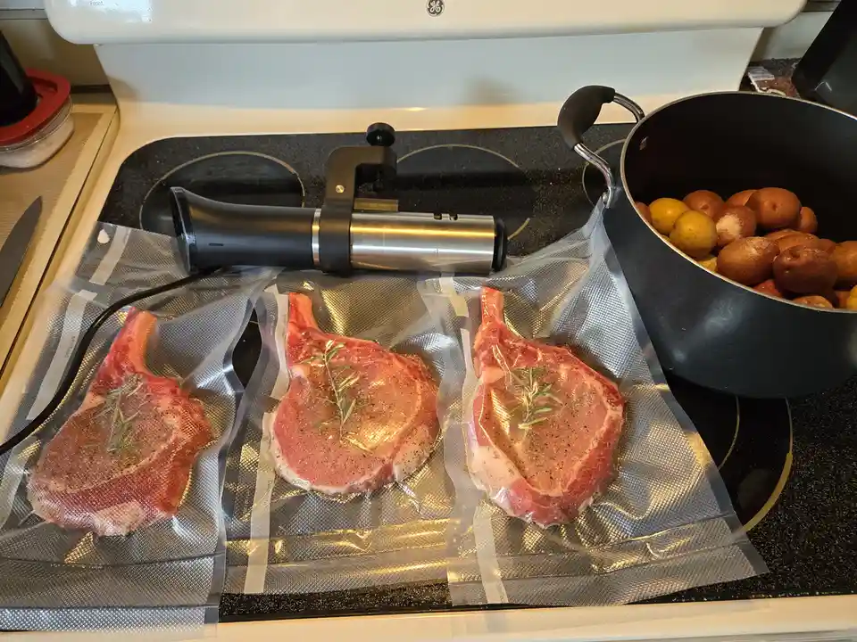 Three raw pork chops, seasoned with rosemary and black pepper, sit in vacuum-sealed bags on a stovetop next to a meat sealer…