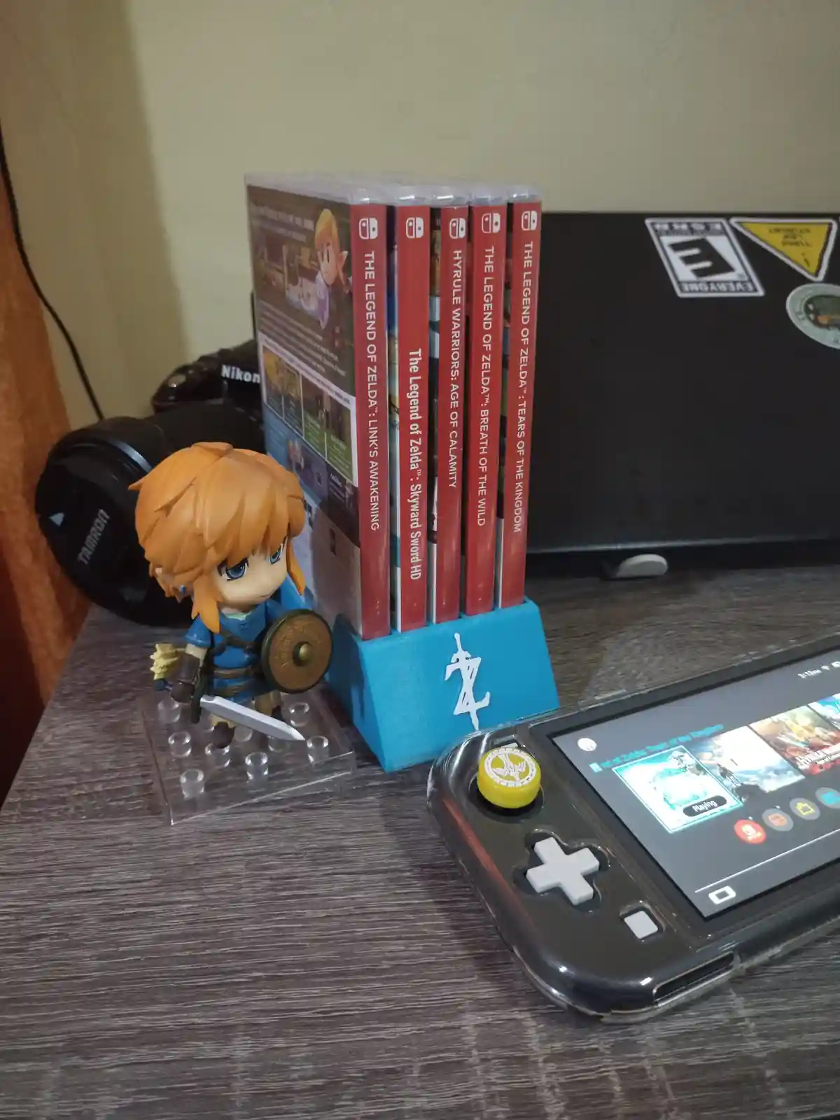 A desk setup featuring a chibi Link figurine, a blue stand holding five Zelda game cases, and a Nintendo Switch Lite playing…