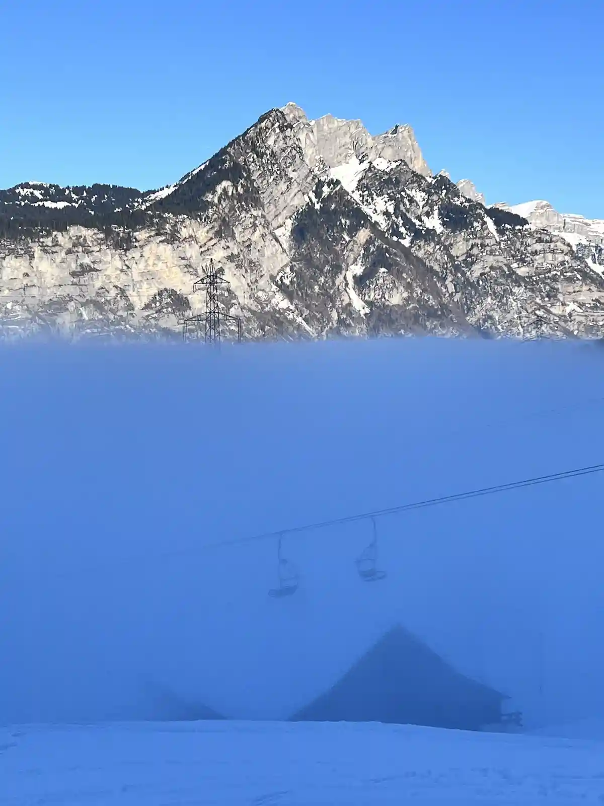 A snowy mountain landscape with a ski lift partially obscured by thick blue fog, with a mountain peak visible above the fog…