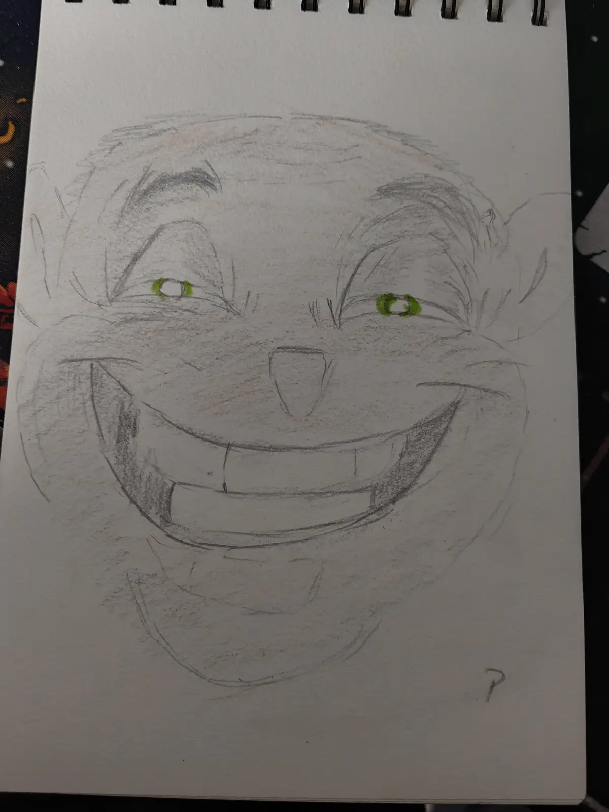 A hand-drawn sketch on a spiral-bound notebook page depicts a cartoonish, wide-grinning face with green eyes and a toothy…