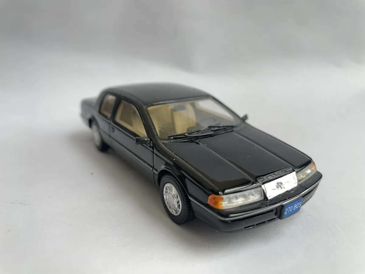 A black die-cast model of a 1980s sedan, likely a Toyota Cressida or similar, is photographed on a plain white surface. The…