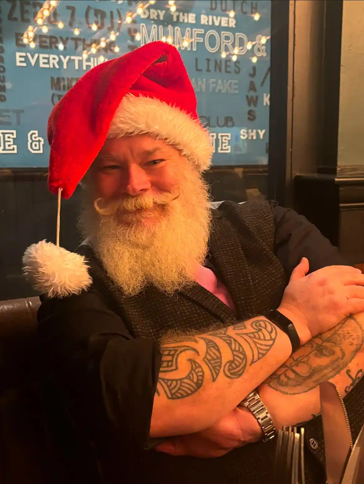 A man with a long white beard and mustache wears a red Santa hat with a white pom-pom while sitting with his arms crossed…