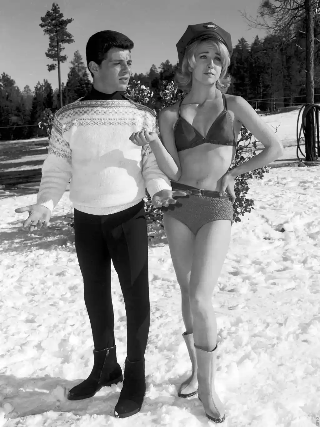 A black-and-white photo shows a man in a patterned sweater and turtleneck standing in snow with his hands out, looking…