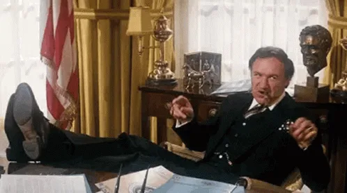 a man in a dark suit reclines with his feet up on a desk in a gilded office, surrounded by American flags and busts. He…