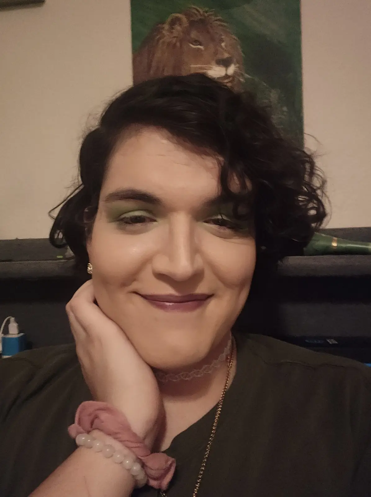 A person with dark curly hair and green eyeshadow smiles while resting their chin on their hand. Behind them, a painting of…