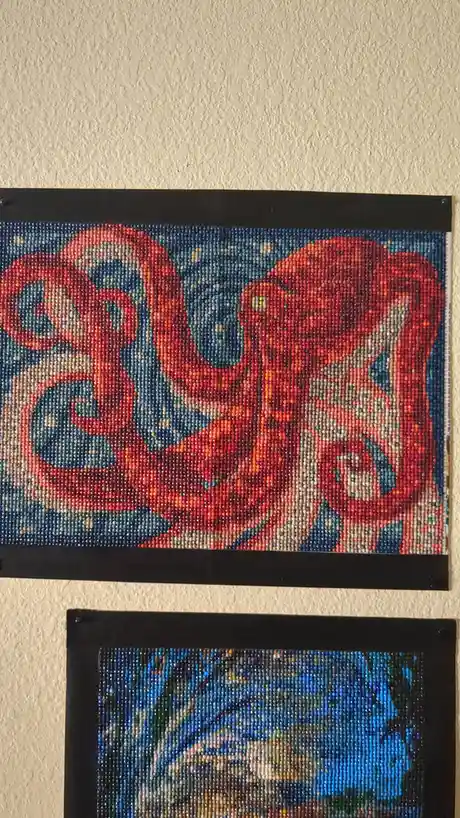 Two framed bead art pieces hang on a textured wall. The top piece is a vibrant red octopus with blue and white beads against…