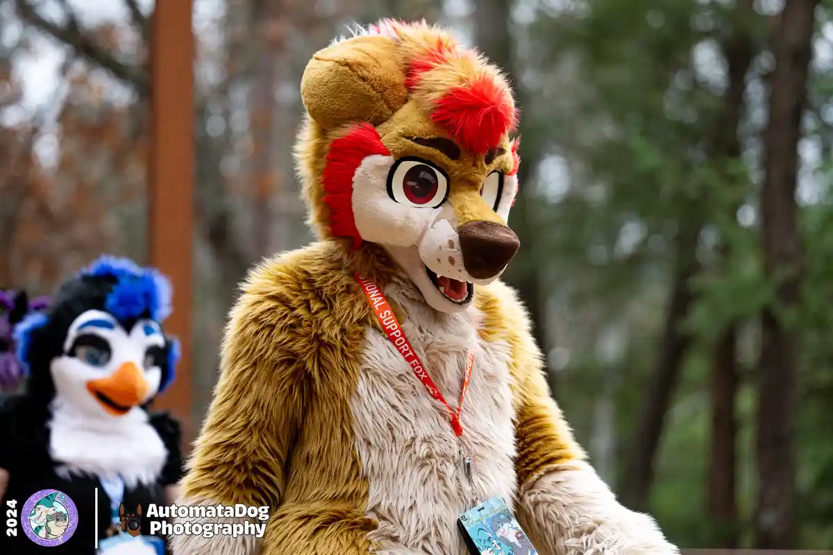 A person in a detailed brown and cream fursuit resembling a fox with red tufts of fur on its head and ears, wearing a red…