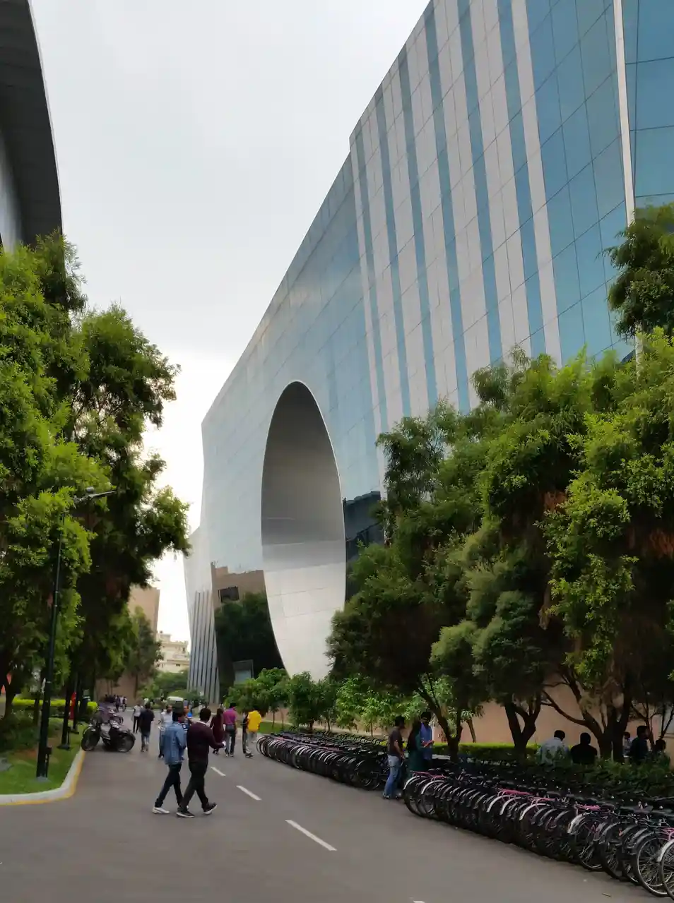 A wide-angle photo shows a modern, curved glass-and-metal building with a large archway, flanked by green trees and rows of…