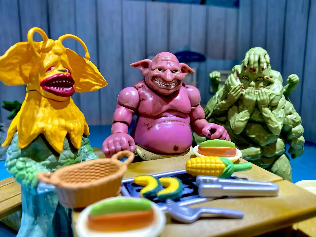Three grotesque, colorful monster action figures sit at a miniature picnic table with toy food. The pink, bald, grinning…