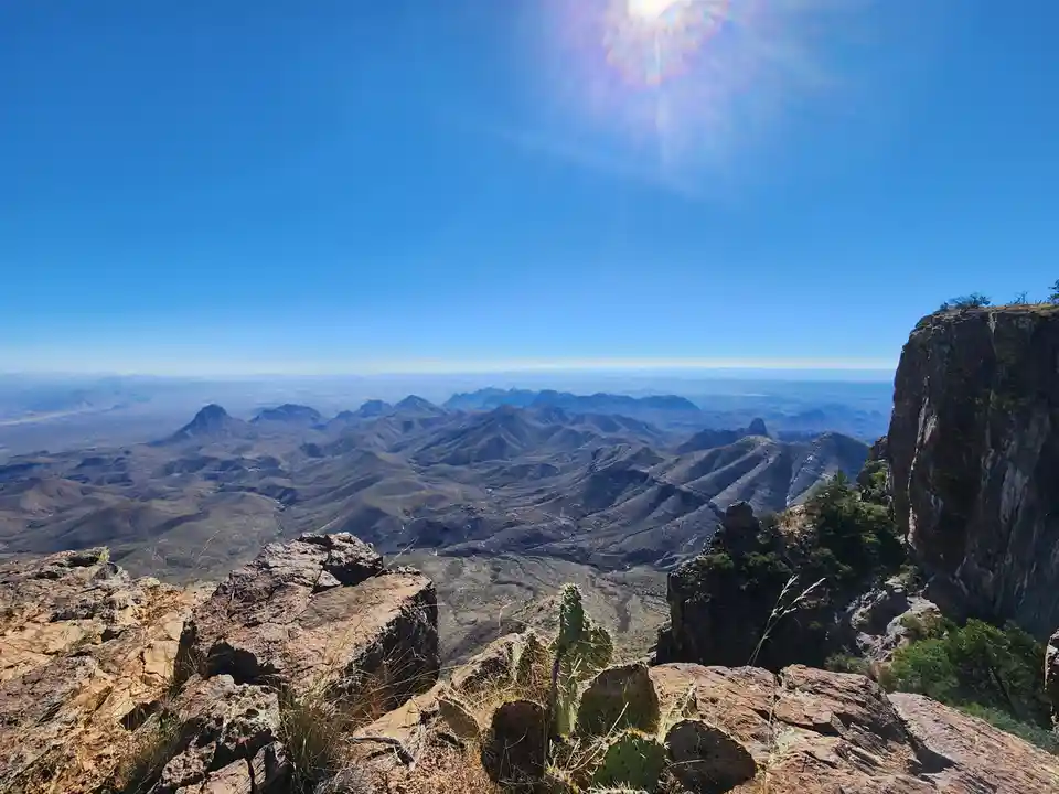 A scenic photo from a high mountain overlook shows a vast desert landscape with rolling hills, distant mountains, and a…