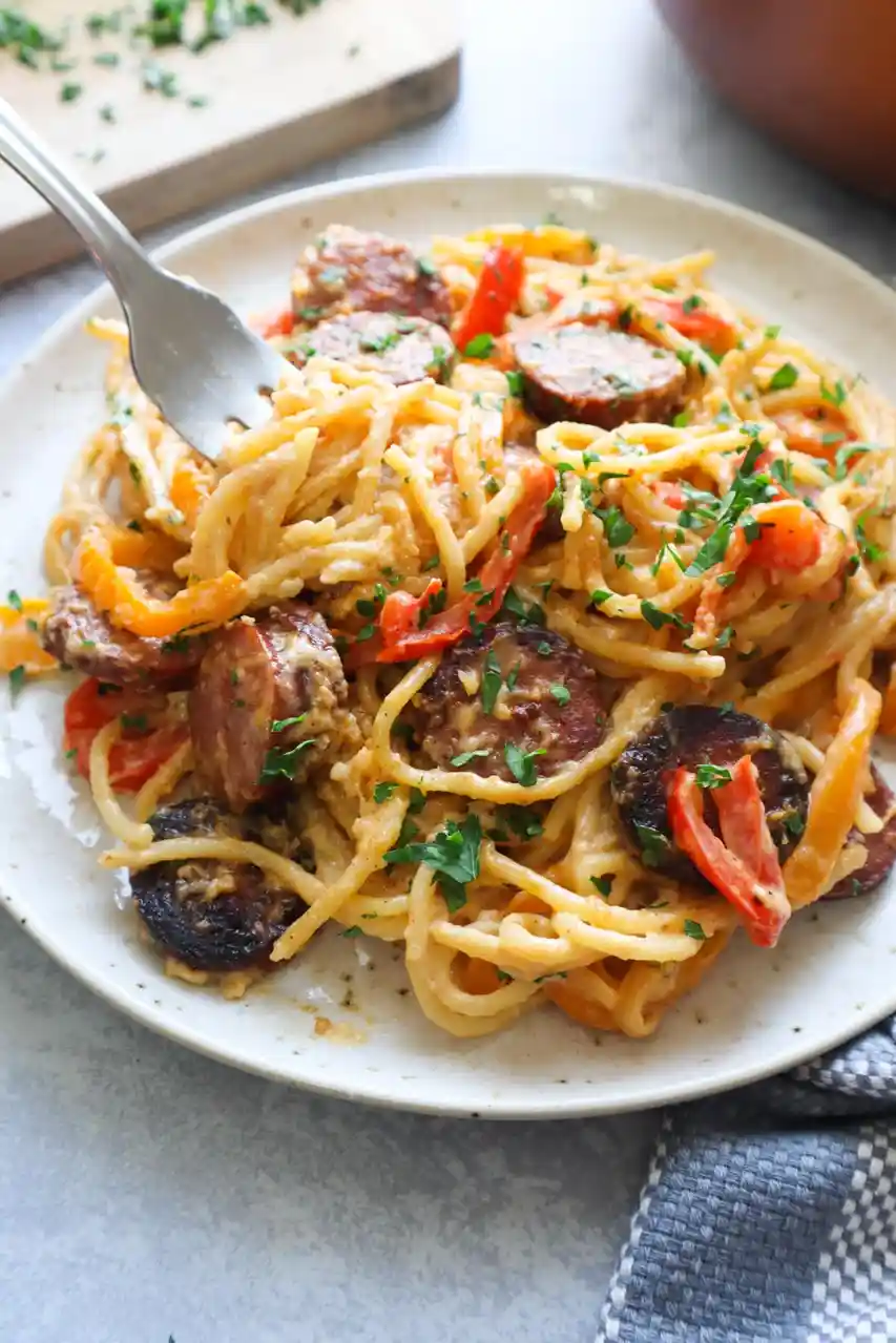 A close-up photo of a plate of creamy pasta mixed with sliced sausage, red and yellow bell peppers, and chopped parsley…