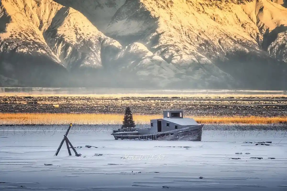 A serene winter landscape shows a frozen lake with snow-covered mountains in the background. A small, abandoned wooden boat…