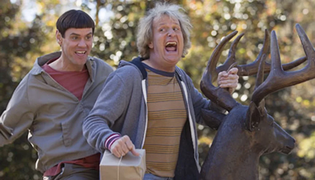 Two men are running outdoors, one holding a wrapped package and the other gripping a large deer statue by its antlers, both…