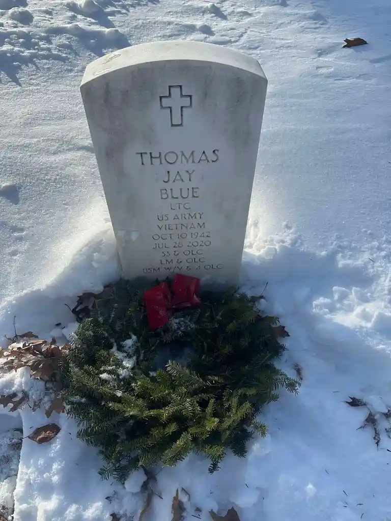 A white military gravestone for Thomas Jay Blue, a U.S. Army LTC who died in 2020, stands in snow with a wreath at its base…