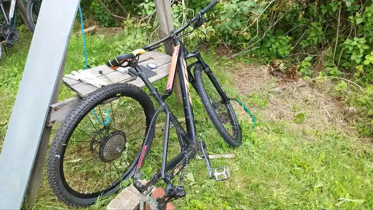 A black mountain bike with orange accents is leaning against a wooden plank and metal beam in a grassy, overgrown outdoor…