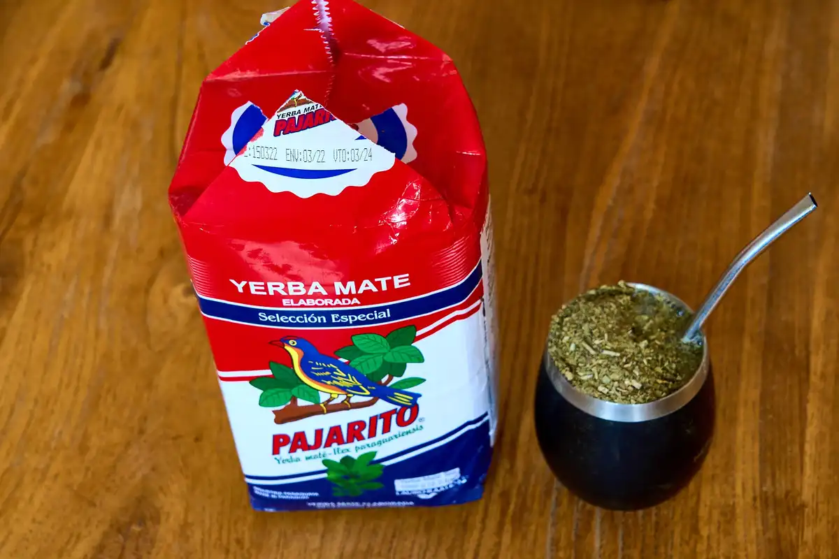 A red and white bag of Pajarito brand yerba mate tea sits next to a traditional black mate gourd filled with dried yerba…