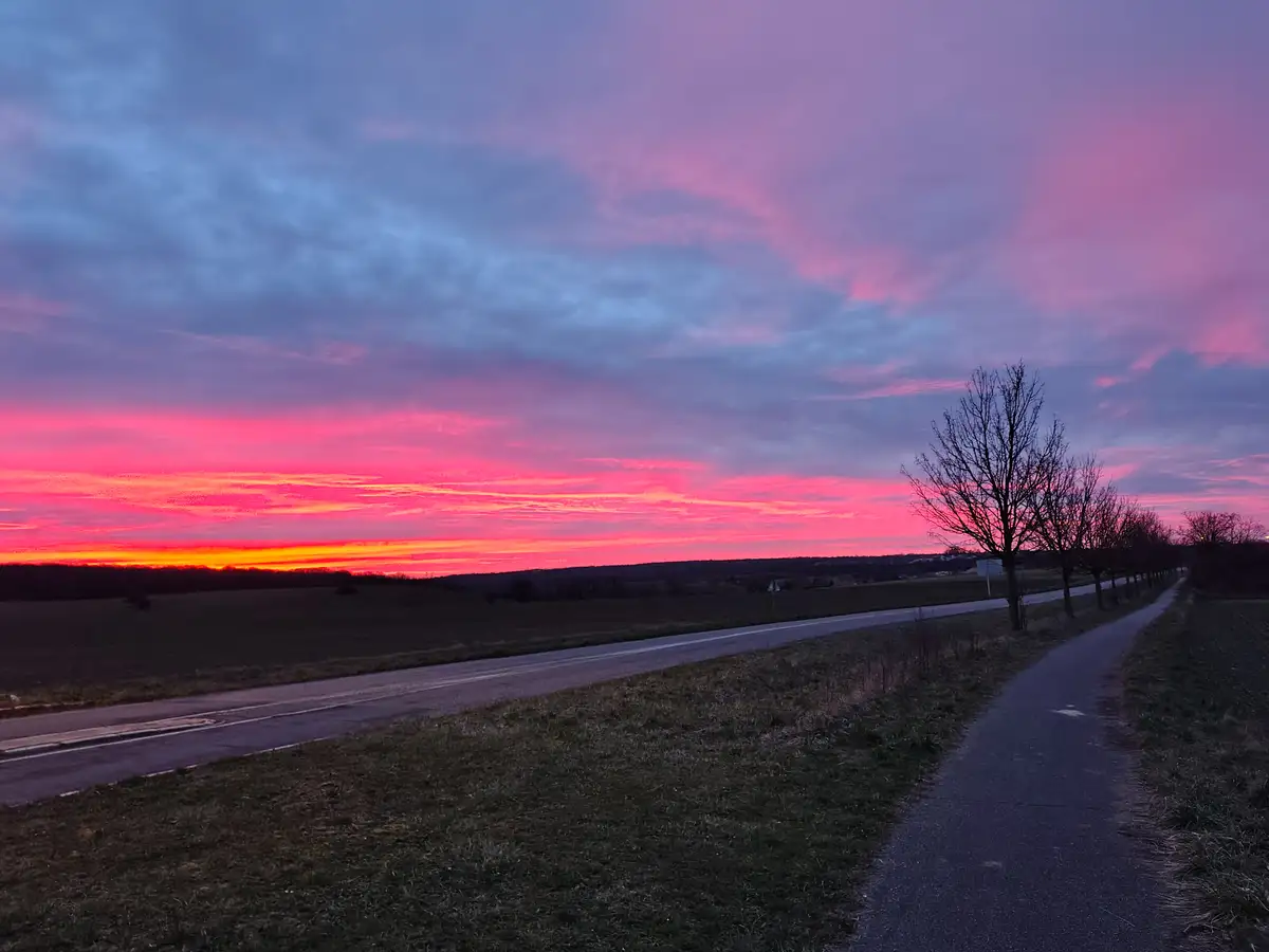 A scenic photo of a rural road at sunset, with vibrant pink, orange, and purple clouds above a dark landscape of fields and…