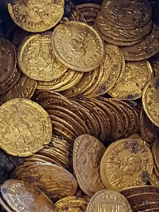 A close-up photo shows a large pile of ancient, tarnished gold coins with embossed portraits and inscriptions, some stacked…