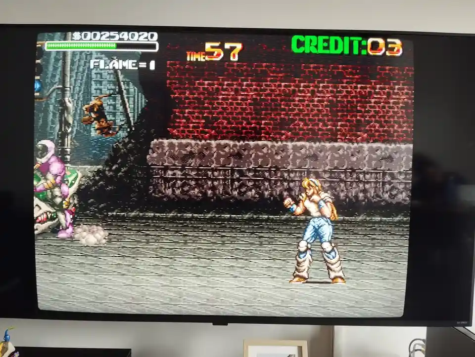 A screenshot of a retro arcade game on a TV screen shows a pixelated character standing in a fighting stance facing a…