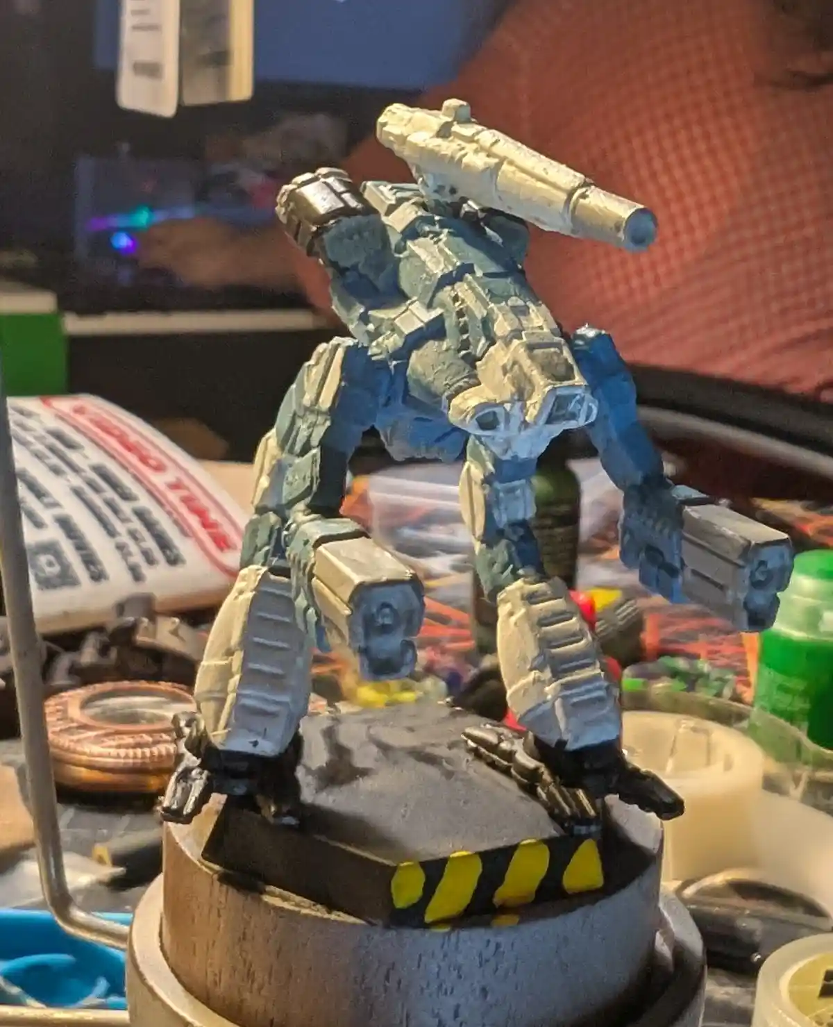 A heavily painted blue and grey robot figurine with multiple weapons stands on a custom base with yellow and black hazard…