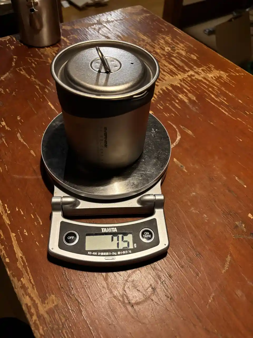 A silver titanium solo pot with a lid sits on a digital kitchen scale that reads '75 g', placed on a worn wooden table. The…