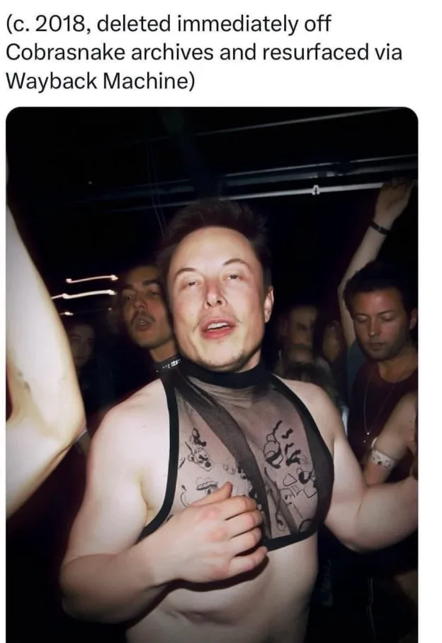 A man with short dark hair and a light beard is wearing a sheer black mesh top over his bare chest, standing in a dimly lit…