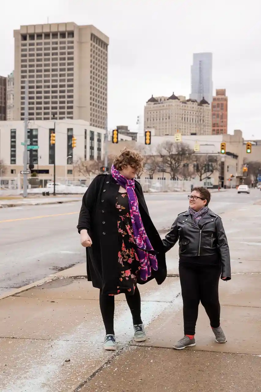 Two people are walking hand-in-hand on a city sidewalk with snow on the ground and tall buildings in the background. The…