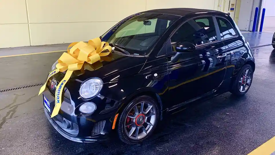 A black Fiat 500 with a large yellow bow tied to its front, featuring 'carmax' written on the bow's ribbon, parked inside a…