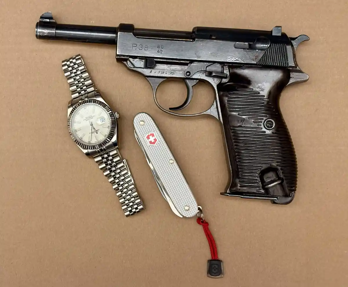 A vintage P.38 pistol is displayed alongside a Rolex Oyster Perpetual Datejust watch and a Swiss Army knife with a red…