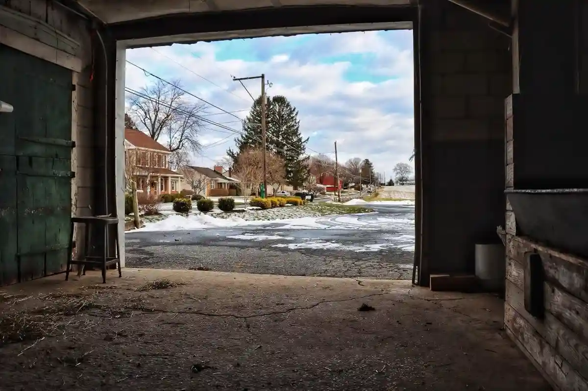 A view from inside a dim, rustic garage looking out onto a snowy suburban street with houses and bare trees under a partly…
