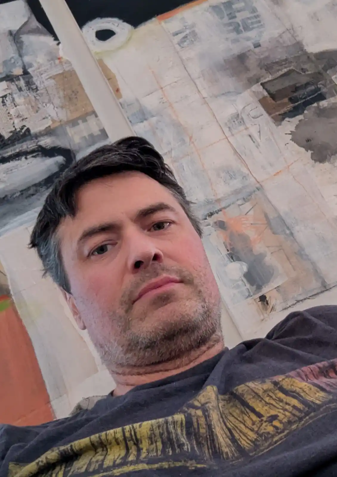 A man with dark hair and a beard looks directly at the camera with a skeptical expression, standing in front of a large…